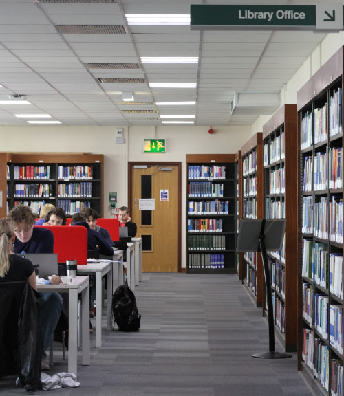 view down an open corridor, on the right hand side wall are book shelves, on the left hand side are rows of students working on desks, at the end of the corridor are more book shelves and a door. 